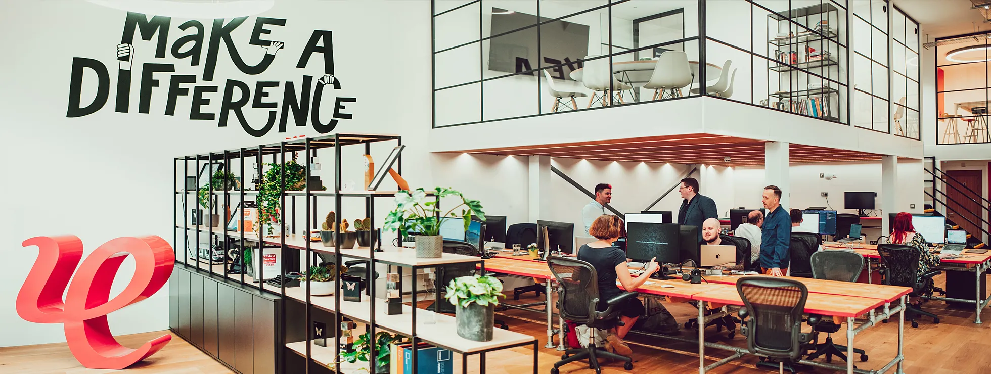 A view across the IE Digital studio in Birmingham, showing members of the agency team collaborating on projects. There's a mural on the wall with the agency's motto "Make a difference, est. 1994" and a large red 3D version of IE's logo marque.  