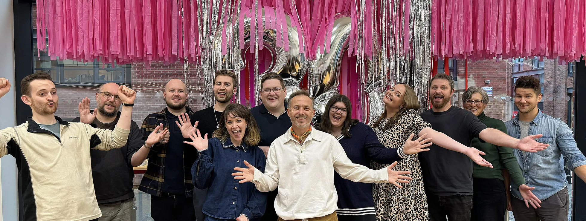 The IE team in the studio celebrating 30 years. Behind them are pink and silver streamers and silver balloons in the shape of the number 30
